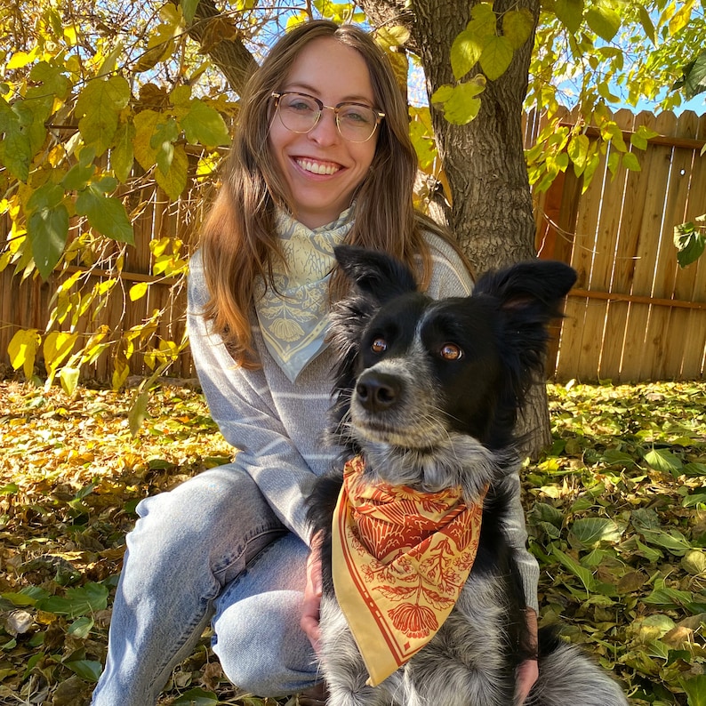 May include: A woman with glasses and a blue bandana is holding a black and white dog with a brown and orange bandana. The dog is wearing a bandana with a floral pattern.