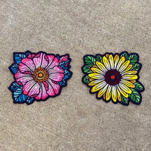 May include: Two embroidered iron-on patches featuring floral designs. One patch is a pink flower with blue and green accents. The other patch is a yellow sunflower with green accents.