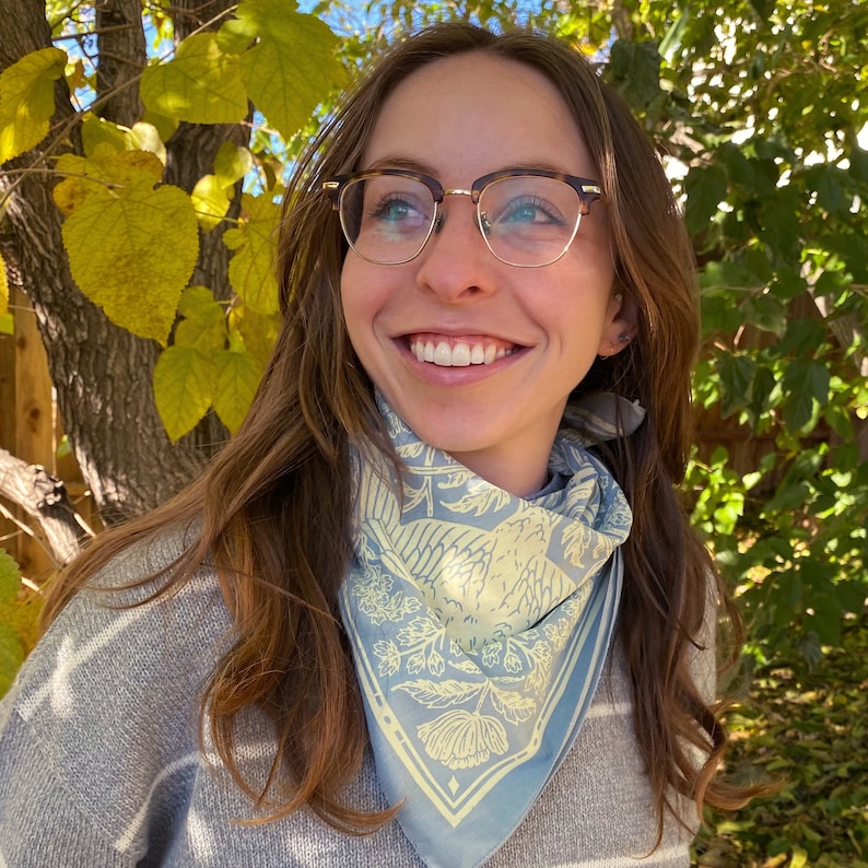 May include: A woman with long brown hair wearing a light blue bandana with a yellow floral and bird design. She is wearing a grey and white striped sweater and brown tortoise shell glasses.
