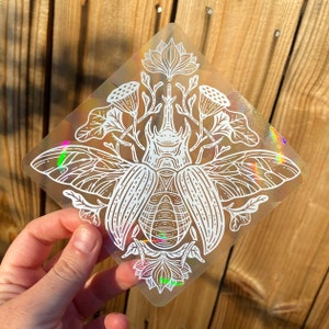 Op de afbeelding: Een witte, iriserende sticker met een gedetailleerde lijntekening van een kever met grote vleugels en een bloemmotief. De kever wordt omringd door bloemen en bladeren.