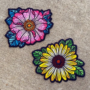 May include: Two embroidered iron-on patches featuring floral designs. The first patch is a pink flower with blue and green leaves. The second patch is a yellow sunflower with green leaves.