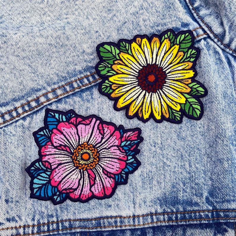 May include: Two embroidered iron-on patches featuring floral designs. One patch is a pink flower with blue and green leaves. The other patch is a yellow sunflower with green leaves.