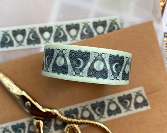 May include: A roll of washi tape with a repeating pattern of moths and crescent moons in shades of green and blue. The tape is on a light green background. A strip of the same tape is visible in the background.