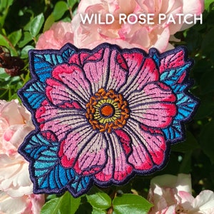 May include: Embroidered patch featuring a pink wild rose with blue and green foliage. The patch has the text "WILD ROSE PATCH" in white lettering.