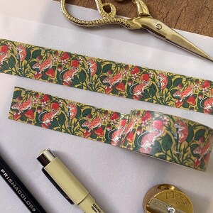 May include: A roll of washi tape with a repeating pattern of red strawberries, green leaves, and pink mice. The tape is on a white surface with a pair of gold scissors and a pencil.
