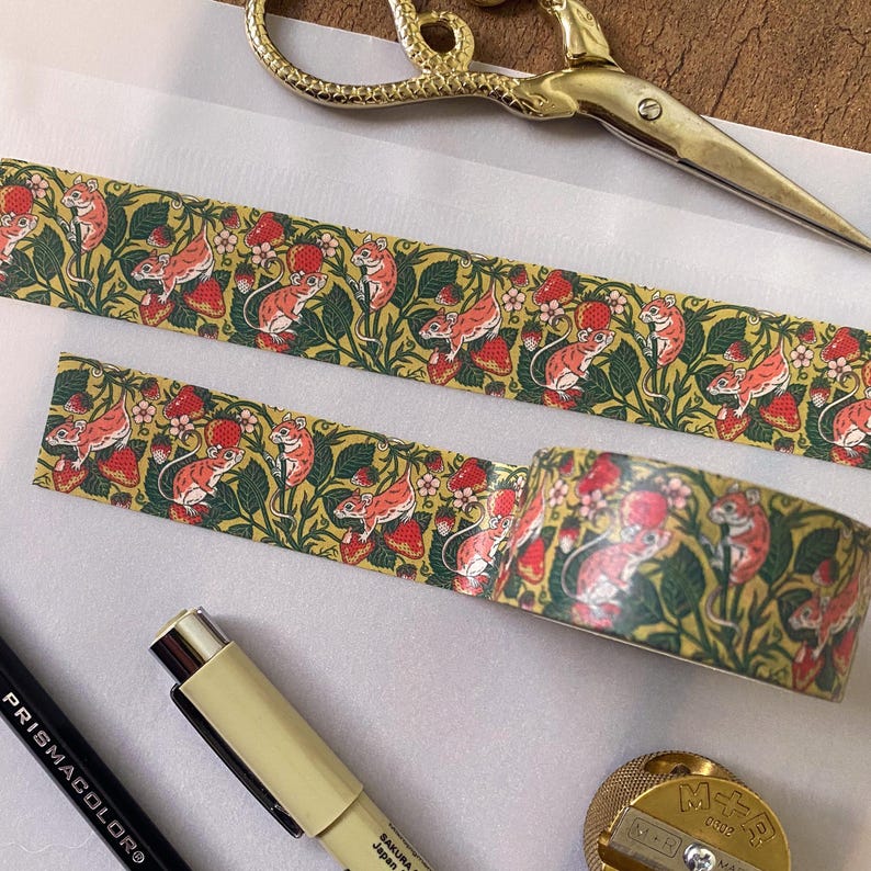 May include: A roll of washi tape with a repeating pattern of red strawberries, green leaves, and pink mice. The tape is on a white surface with a pair of gold scissors and a pencil.