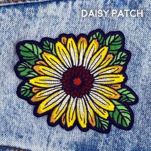 May include: A yellow and white embroidered daisy flower patch with green leaves. The patch has the text "DAISY PATCH" in black letters.