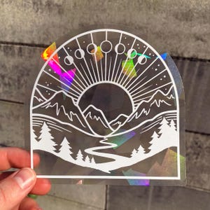 Könnte beinhalten: Ein weißer Vinyl-Aufkleber mit holografischem Hintergrund, der eine Bergkette, einen Fluss, eine Sonne und eine zunehmende Mondsichel mit Phasen zeigt. Das Design hat die Form eines Halbkreises.
