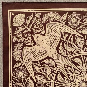 May include: A brown fabric bandana with a white bird and floral design. The bird is flying with its wings spread and is surrounded by flowers and leaves.