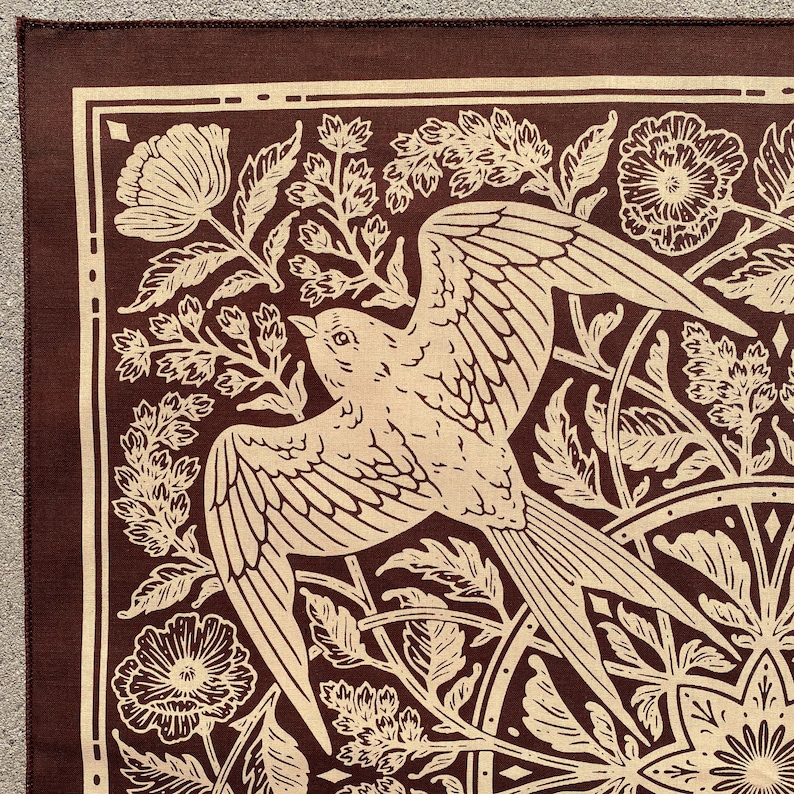 May include: A brown fabric bandana with a white bird and floral design. The bird is flying with its wings spread and is surrounded by flowers and leaves.