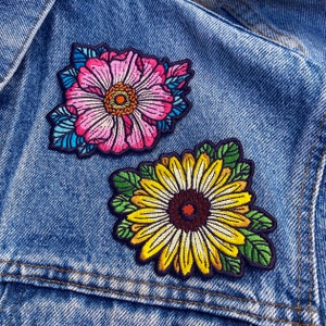 May include: Two embroidered iron-on patches featuring floral designs. The first patch is a pink flower with blue and green accents. The second patch is a yellow sunflower with green leaves.