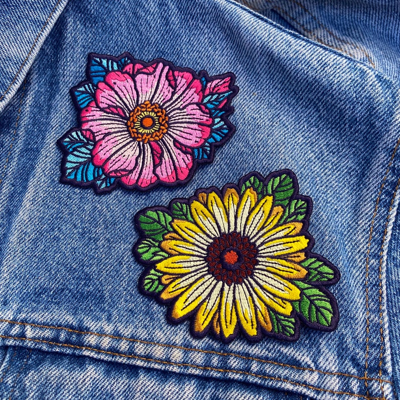 May include: Two embroidered iron-on patches featuring floral designs. The first patch is a pink flower with blue and green accents. The second patch is a yellow sunflower with green leaves.
