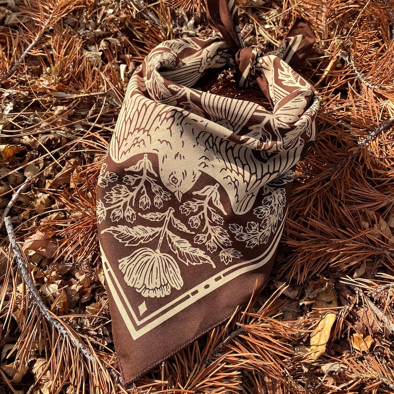 May include: A brown bandana with a white floral and bird design. The bandana is folded and tied in a knot.