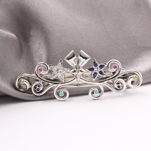 May include: A silver hair barrette with a decorative floral design. The barrette features two star-shaped flower accents, one with clear stones and the other with blue stones. Small, colourful gemstones are set throughout the design.