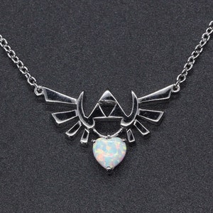 May include: Silver-toned necklace featuring a stylized wing design with a central triangle and a heart-shaped opal pendant. The necklace has a delicate chain and is set against a dark background.