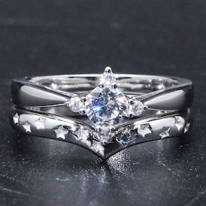 Op de afbeelding: Zilveren verlovingsring set met een solitaire diamant en een met sterren bezaaide band. De verlovingsring heeft een centrale, grote, heldere diamant met kleinere accentstenen. De band heeft een V-vorm en is versierd met kleine stervormige uitsparingen.