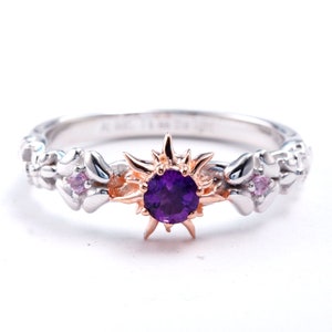 May include: A silver ring with a central purple gemstone, framed by a gold-colored sunburst design. The band features floral and pink accents. A unique piece of jewelry.