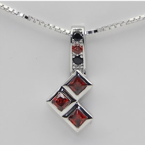 Może przedstawiać: Srebrny naszyjnik z wisiorkiem o geometrycznym wzorze. Wisiorek ma czerwone i czarne kamienie szlachetne, z prostokątną górą i dwoma kwadratowymi czerwonymi kamieniami. Łańcuszek naszyjnika jest delikatny, srebrny.