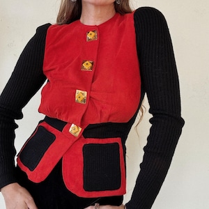 May include: A red and black sweater with gold buttons. The sweater has a unique design with a red suede panel in the front and black knit sleeves and bottom.