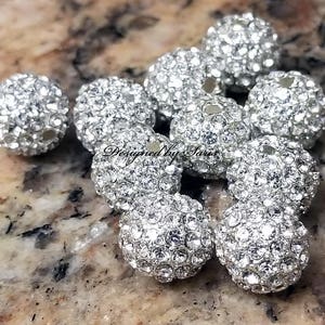 Puede incluir: Un conjunto de cuentas brillantes de estr&aacute;s plateadas. Cada cuenta redonda est&aacute; cubierta de peque&ntilde;os cristales reflectantes. El texto "Designed By Saria" es visible en las cuentas. Ideal para la creaci&oacute;n de joyas y diversos proyectos de artesan&iacute;a.