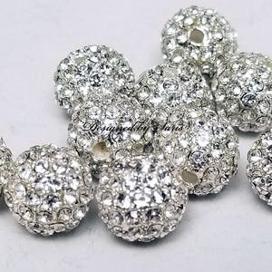 Puede incluir: Un conjunto de cuentas redondas de color plateado incrustadas con peque&ntilde;os y brillantes pedrer&iacute;a. Las cuentas est&aacute;n dispersas sobre una superficie blanca, con el texto "Designed by Janis" visible en una cuenta.