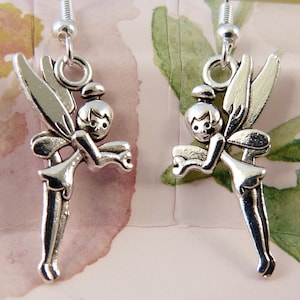 May include: Silver earrings featuring a fairy design with wings and a small face. The fairy is holding a small object in its hands.