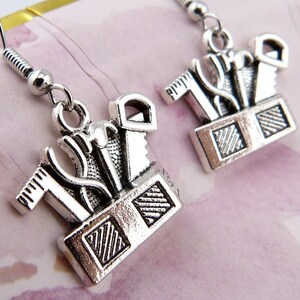 May include: Silver tone earrings featuring a toolbox design with various tools, including a hammer, saw, and wrench. The tools are arranged in a small toolbox with a textured surface.