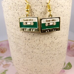 May include: A pair of gold-tone earrings shaped like cassette tapes with green and white enamel. The earrings have a spring-loaded hook closure.