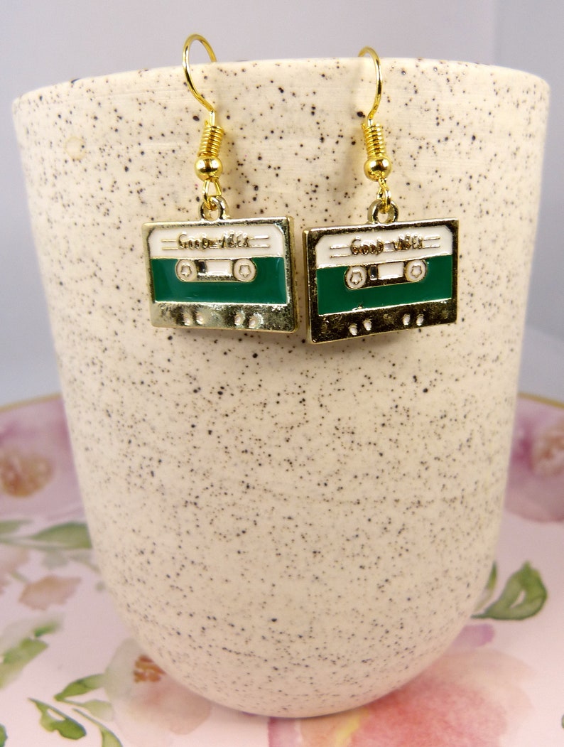 May include: A pair of gold-tone earrings shaped like cassette tapes with green and white enamel. The earrings have a spring-loaded hook closure.
