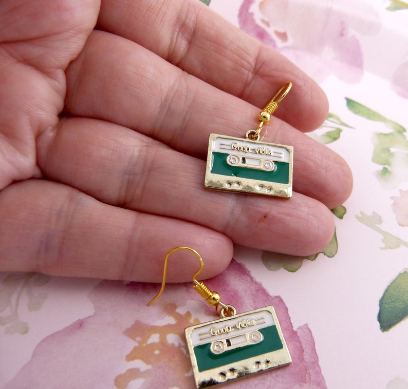 May include: A pair of gold tone earrings with green and white cassette tape charms. The charms have the text "Good Vibes" on them.