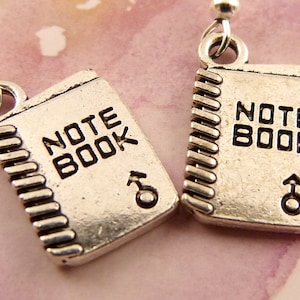 May include: A pair of silver earrings shaped like a notebook with the word "NOTEBOOK" engraved on the front. Each earring has a small silver charm in the shape of a male symbol.