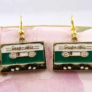 May include: A pair of gold-tone earrings with a green and white cassette tape design. The tape has the words "Good Vibes" printed on it.