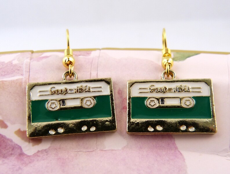 May include: A pair of gold-tone earrings with a green and white cassette tape design. The tape has the words "Good Vibes" printed on it.