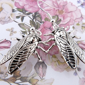 May include: A pair of silver earrings featuring detailed cicada insect designs. The earrings have a dangling style with a hook fastening.