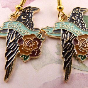 May include: A pair of gold-tone earrings featuring black enamel crows with a rose and a banner that reads "Solitude".