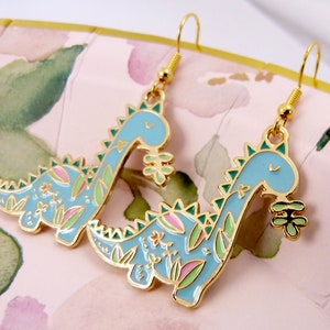 K&ouml;nnte beinhalten: Ein Paar goldfarbene Ohrh&auml;nger mit einem blauen Dinosaurier-Design mit gr&uuml;nen und rosa floralen Akzenten. Die Dinosaurier halten eine kleine gr&uuml;ne Blume.