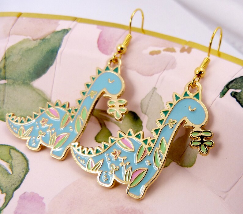 K&ouml;nnte beinhalten: Ein Paar goldfarbene Ohrh&auml;nger mit einem blauen Dinosaurier-Design mit gr&uuml;nen und rosa floralen Akzenten. Die Dinosaurier halten eine kleine gr&uuml;ne Blume.