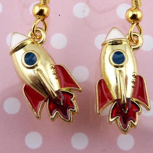 May include: A pair of gold rocket ship earrings with red and white enamel details. The rockets have a blue enamel circle for the window.