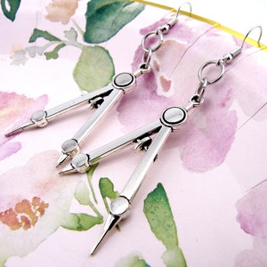 May include: A pair of silver compass earrings. The earrings are in the shape of a compass with a pointed tip and a circle at the top. The earrings are hanging from silver hooks.