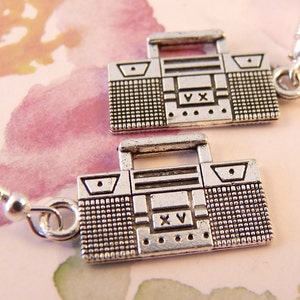 May include: Silver earrings featuring a boombox design. The boomboxes have a silver finish and a textured surface. The earrings are suspended from silver hooks.