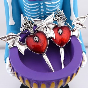 May include: A pair of silver earrings with red heart charms and bat wings. The hearts have a skull design and are accented with a metallic finish.