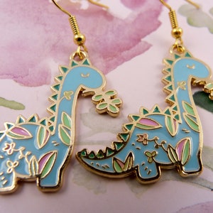 K&ouml;nnte beinhalten: Ein Paar goldfarbene Ohrringe mit einem blauen Dinosaurier-Design mit gr&uuml;nen und rosa floralen Akzenten.