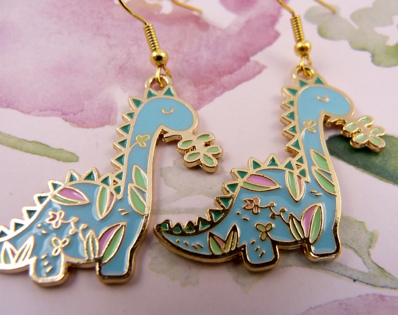 K&ouml;nnte beinhalten: Ein Paar goldfarbene Ohrringe mit einem blauen Dinosaurier-Design mit gr&uuml;nen und rosa floralen Akzenten.