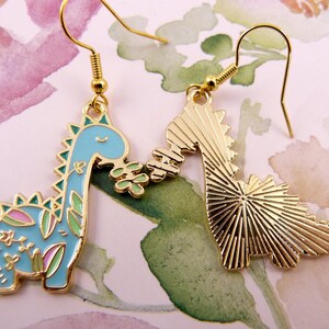 K&ouml;nnte beinhalten: Ein Paar goldfarbene Ohrringe mit einem blauen und gr&uuml;nen Dinosaurier mit floralen Details auf der einen Seite und einem massiven goldenen Dinosaurier auf der anderen Seite.