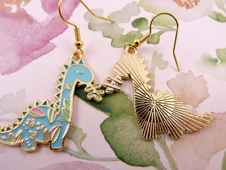 K&ouml;nnte beinhalten: Ein Paar goldfarbene Ohrringe mit einem blauen und gr&uuml;nen Dinosaurier mit floralen Details auf der einen Seite und einem massiven goldenen Dinosaurier auf der anderen Seite.