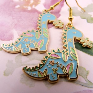 K&ouml;nnte beinhalten: Ein Paar goldfarbene Ohrringe mit einem blauen Dinosaurier-Design mit gr&uuml;nen und rosa floralen Akzenten. Die Dinosaurier sind in entgegengesetzte Richtungen gerichtet.