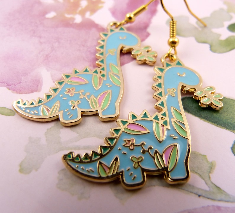 K&ouml;nnte beinhalten: Ein Paar goldfarbene Ohrringe mit einem blauen Dinosaurier-Design mit gr&uuml;nen und rosa floralen Akzenten. Die Dinosaurier sind in entgegengesetzte Richtungen gerichtet.