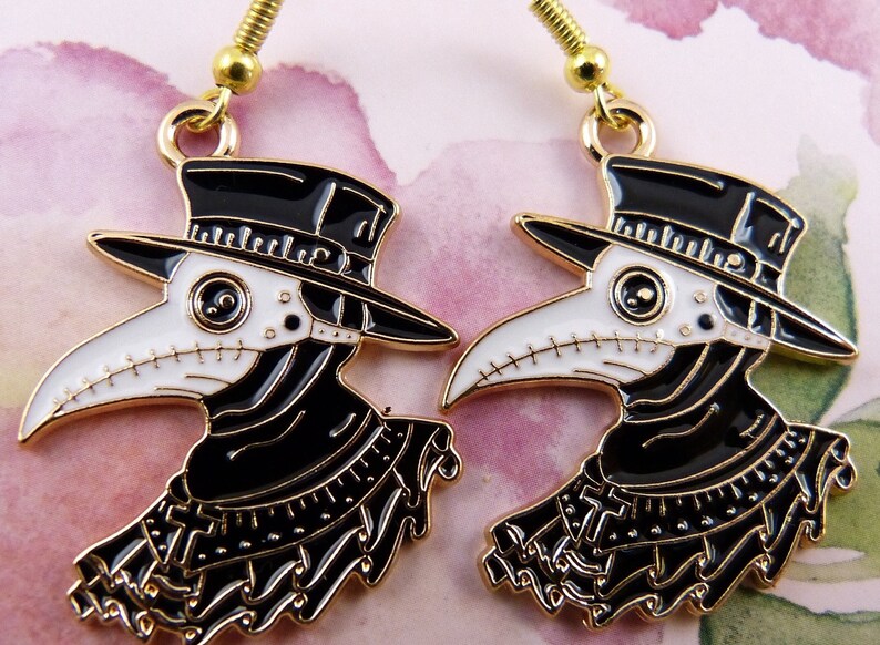 Puede incluir: Un par de pendientes de tono dorado con un dise&ntilde;o de m&eacute;dico de la peste en blanco y negro. Los pendientes est&aacute;n hechos de metal y tienen un sombrero negro, una m&aacute;scara blanca y ribetes negros y dorados.