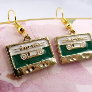 May include: Gold tone earrings with a green and white cassette tape charm. The charm has the words "Good Vibes" written on it.