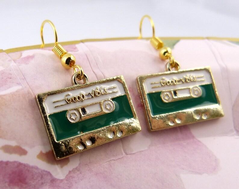 May include: Gold tone earrings with a green and white cassette tape charm. The charm has the words "Good Vibes" written on it.